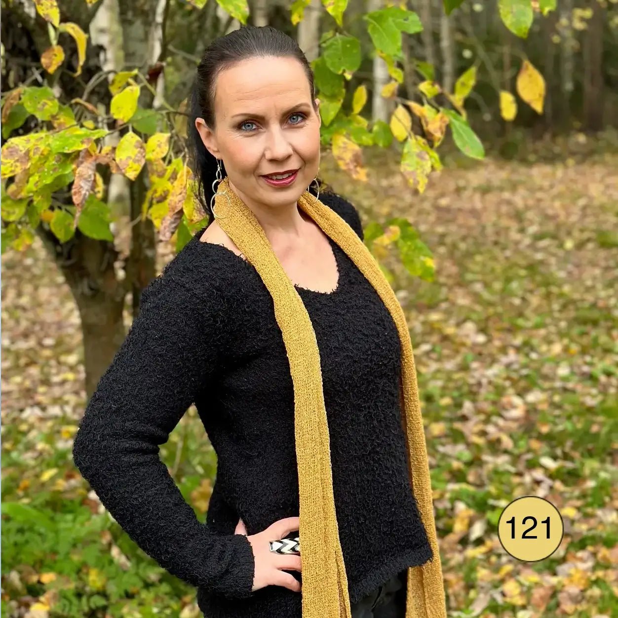 Mörkhårig kvinna utomhus i snö med en Almira sjal i ljus senap virad runt halsen över en svart tröja; den långa stickade sjalen lyser varmt gulbrun mot det vita. Ulla Jacobsson Kläder
