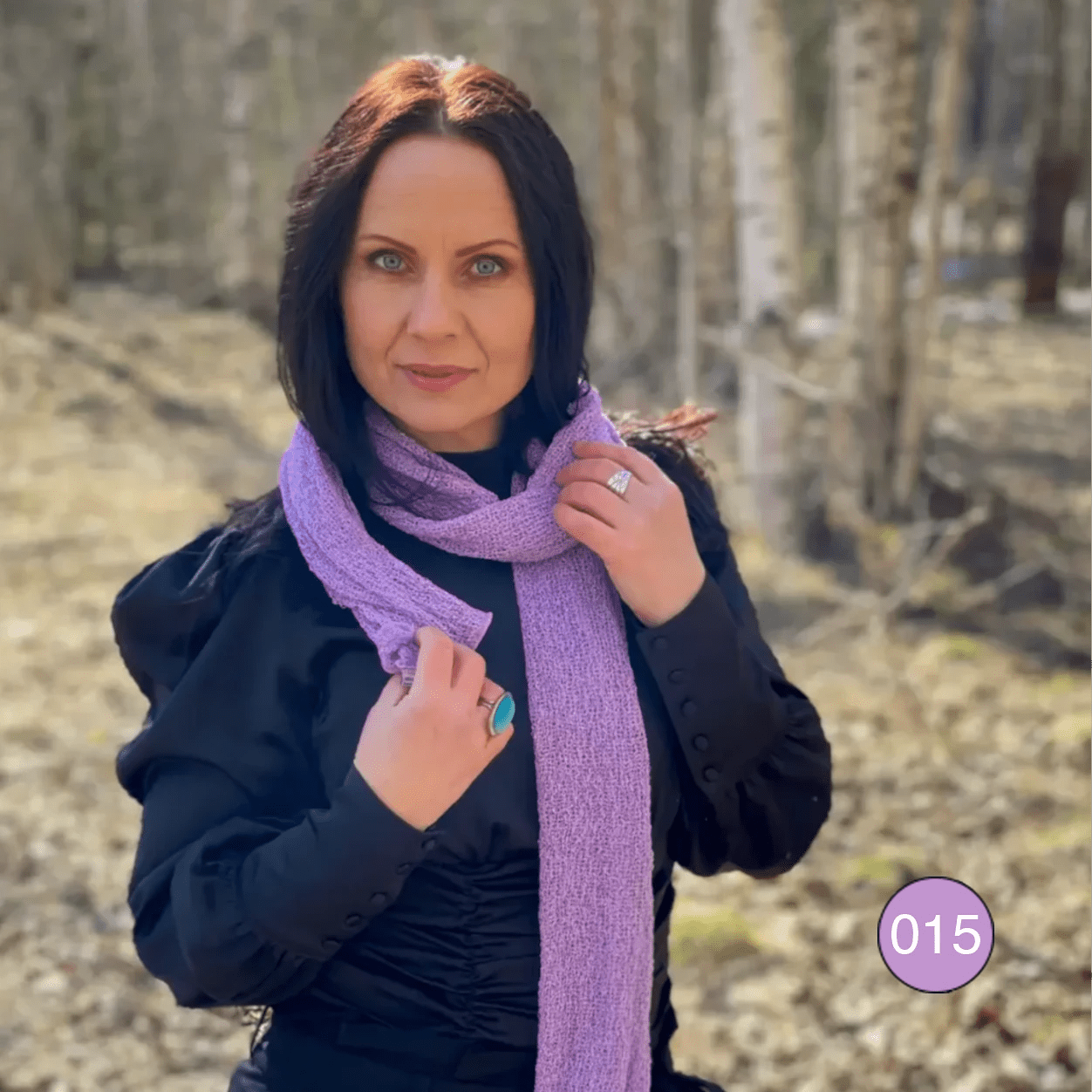 Kvinna som håller en Almira sjal i lupinlila kring axlarna med händerna nära kragen; det finstickade tyget lägger sig i mjuka veck mot en mörk bakgrund. Ulla Jacobsson Kläder