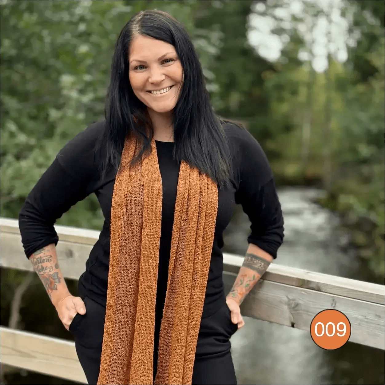 Almira sjal i mocka virad runt halsen på en kvinna vid ett räcke nära vatten; den långa bruna sjalen hänger rakt ned över en svart tröja och visar garnets matta yta. Ulla Jacobsson Kläder