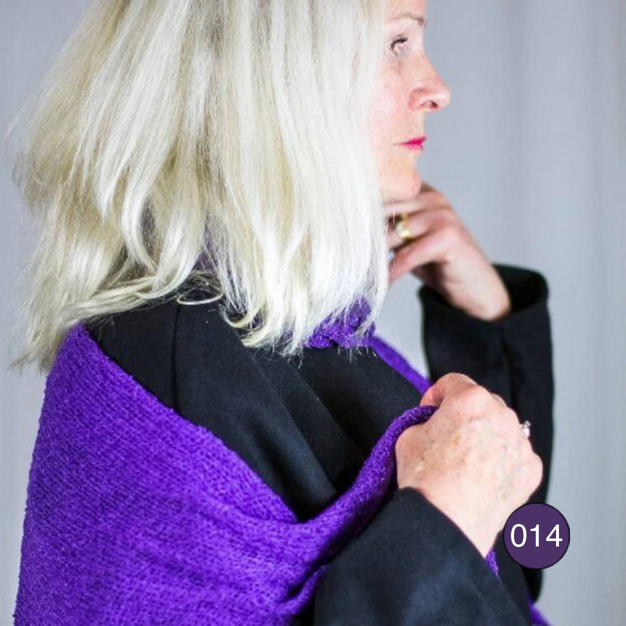 Ljushårig kvinna med en Almira sjal i mörklila draperad runt halsen, sedd snett från sidan; det stickade garnet vilar i mjuka veck mot kinden. Ulla Jacobsson Kläder