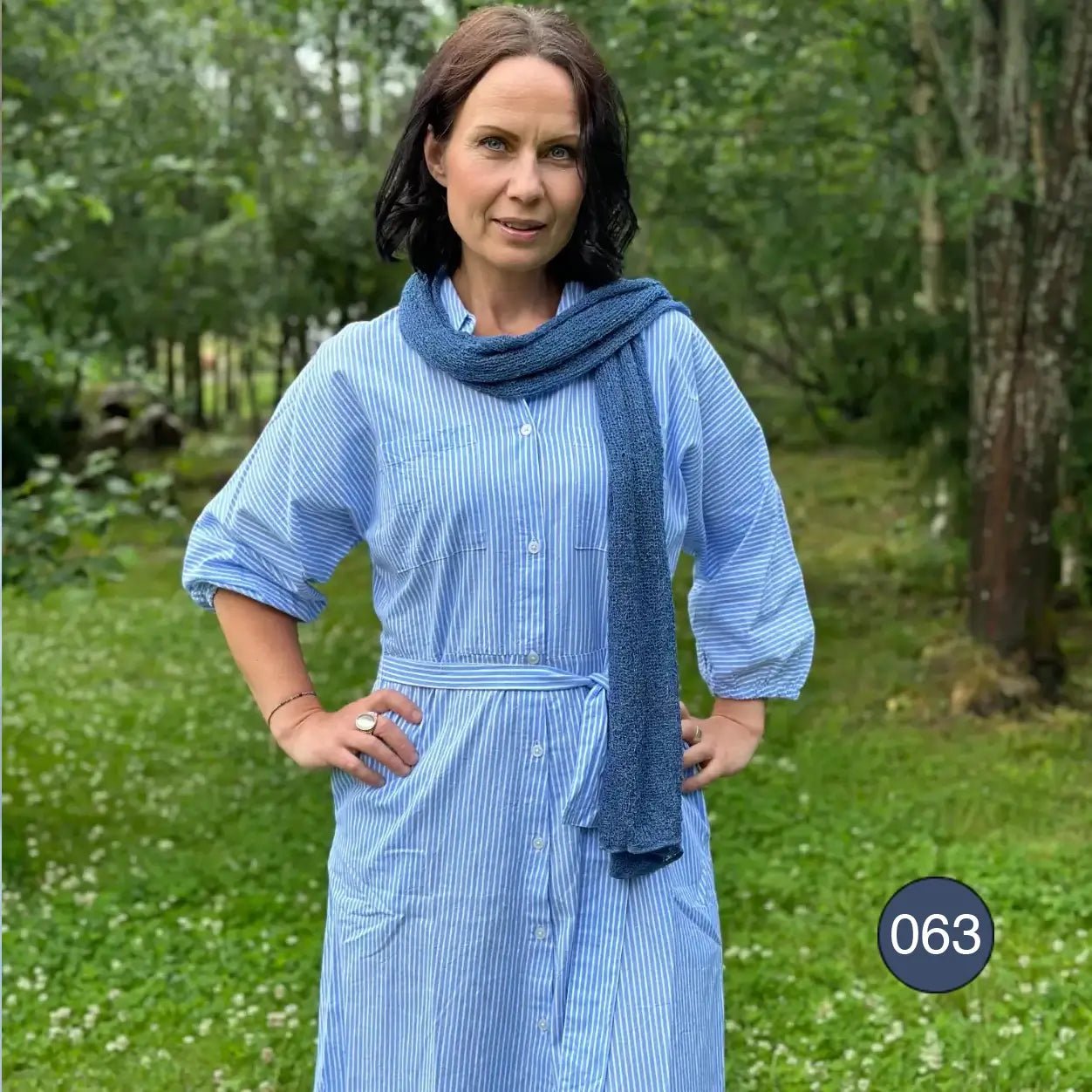 Mörkhårig kvinna utomhus i grönska, insvept i en stor Almira sjal i ocean-blått som läggs som ett mjukt sjok över axlar och rygg. Ulla Jacobsson Kläder