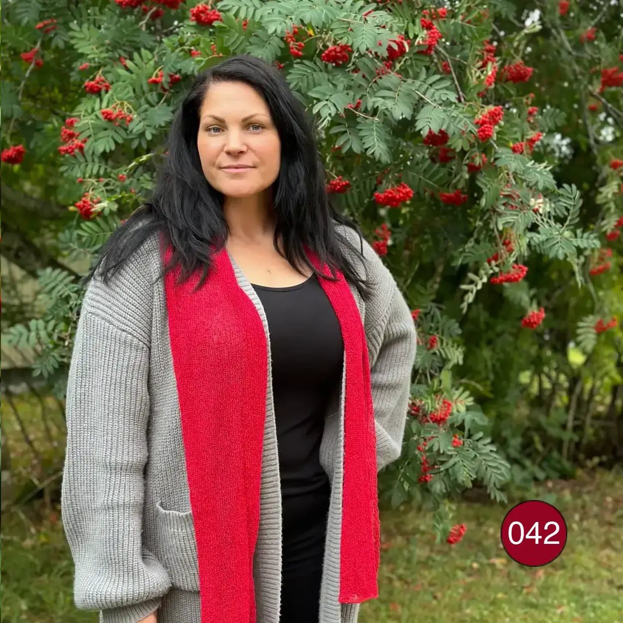 Almira sjal i röd ton som hänger långt nedför framsidan på en kvinna i svart, utomhus intill en rönn med röda bär; den stickade sjalen faller mjukt i raka längder. Ulla Jacobsson Kläder