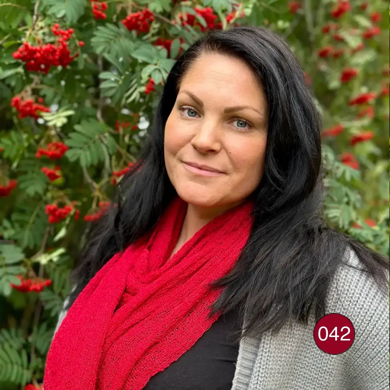 Mörkhårig kvinna utomhus bland röda bär, med en Almira sjal i röd ton draperad runt halsen och nedhängande längder mot en svart tröja. Ulla Jacobsson Kläder