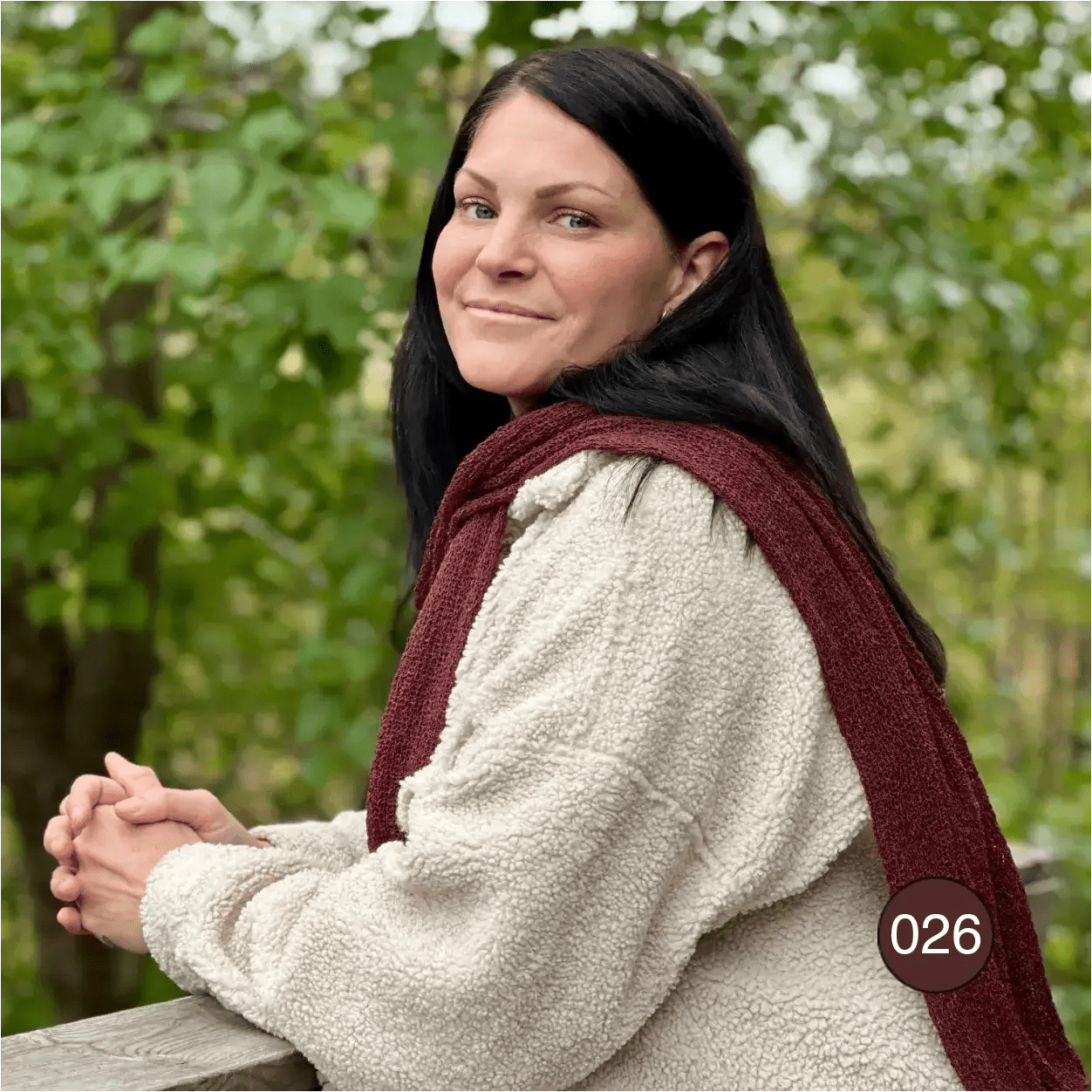 Kvinna i grovstickad cremefärgad tröja med en rödbrun Almira sjal löst draperad runt halsen; det finmaskiga garnet kontrasterar mot den kraftiga stickningen. Ulla Jacobsson Kläder