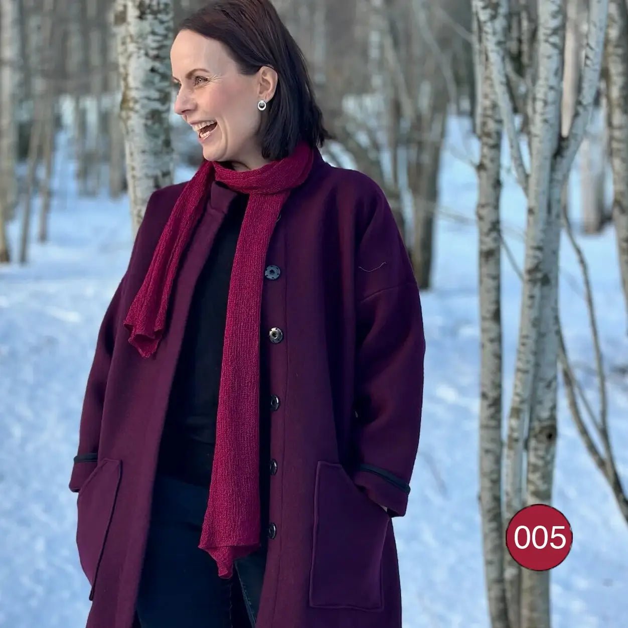 Almira sjal i vinröd ton lagd i dubbla varv runt halsen på en mörkhårig kvinna utomhus; den långa stickade sjalen ramar in ansiktet och faller ned över en mörk tröja. Ulla Jacobsson Kläder