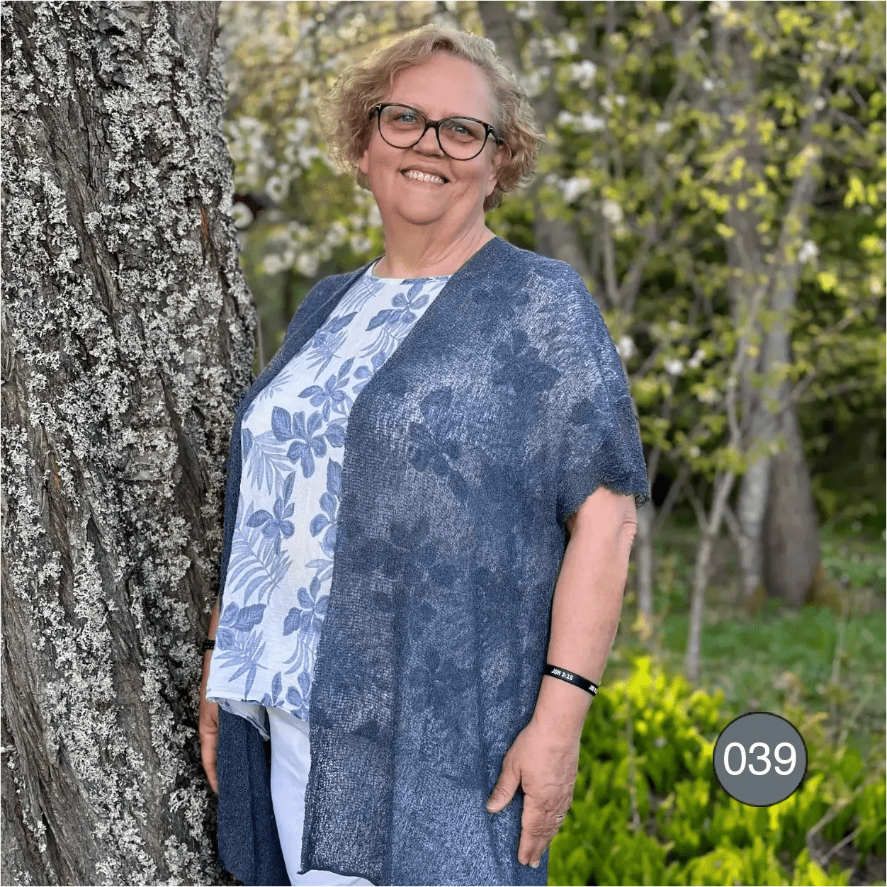 Äldre kvinna med glasögon i en Biasa kofta i anilin, en ljus blåmelerad ton; det finstickade garnet är luftigt och faller öppet över ett vitt plagg. Ulla Jacobsson Kläder