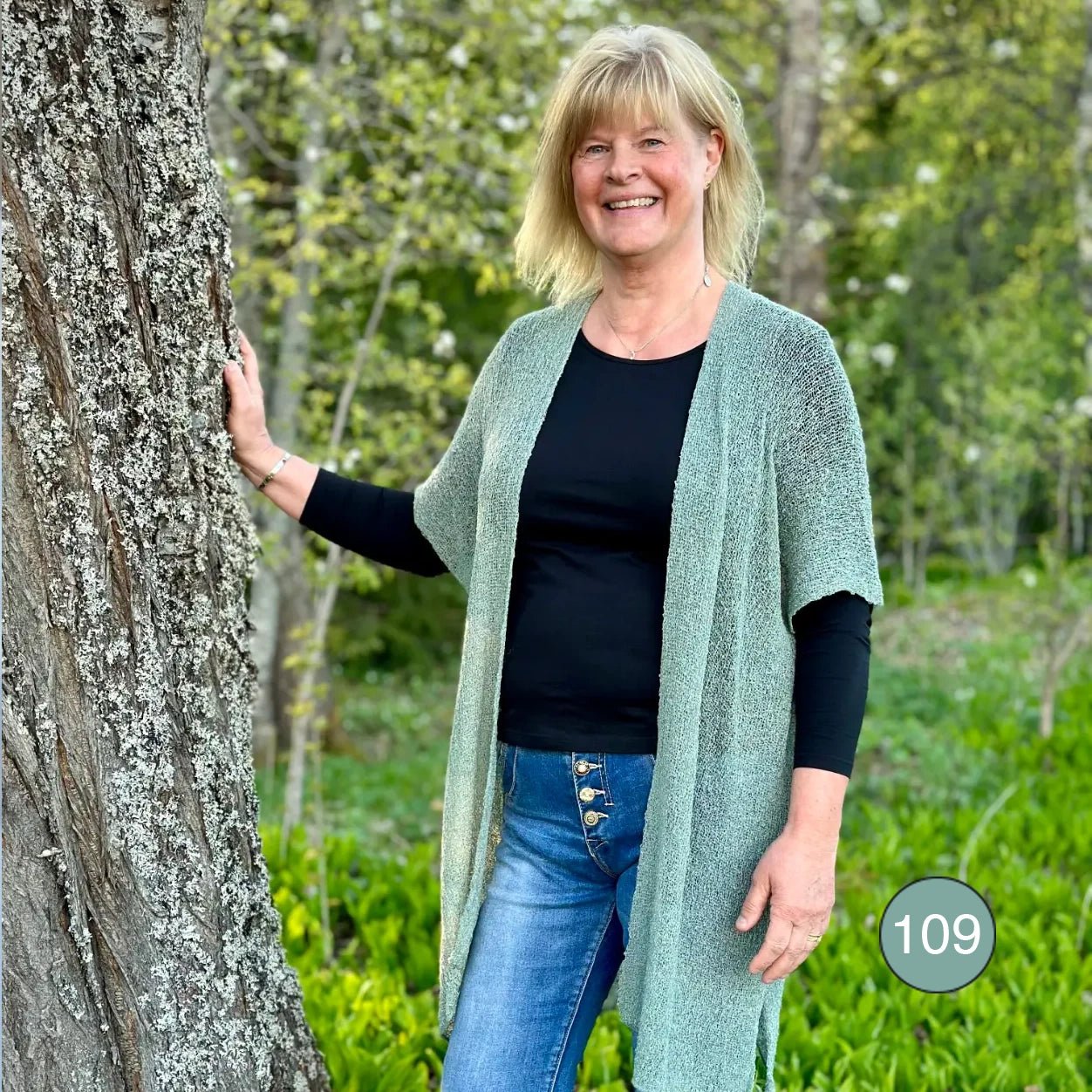 Ljushårig kvinna med handen mot en trädstam i en Biasa kofta i grå mossa, en dämpad grågrön ton; den långa stickningen hänger öppen över svart och jeans. Ulla Jacobsson Kläder