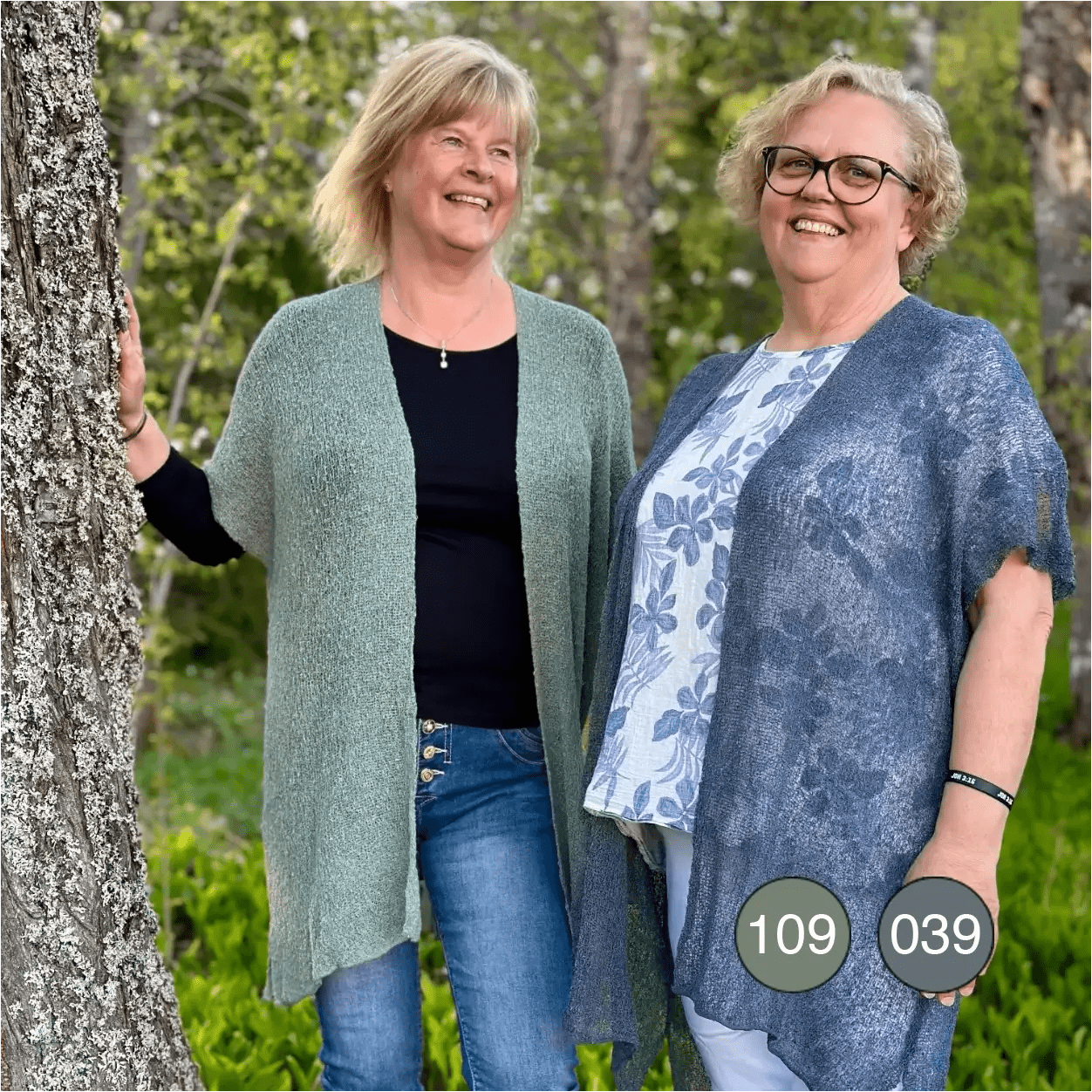 Två kvinnor utomhus, den ena i en Biasa kofta i grå mossa och den andra i anilin blåmelerad; båda långa och finstickade, öppna över mörka plagg och jeans. Ulla Jacobsson Kläder