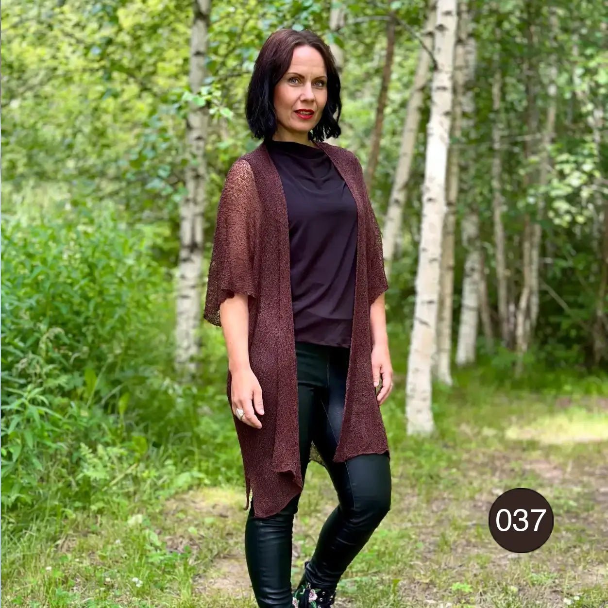 Mörkhårig kvinna utomhus i grönska i en mörkbrun Biasa kofta över en brun polotröja; den långa finstickade koftan faller mjukt nedför kroppen. Ulla Jacobsson Kläder