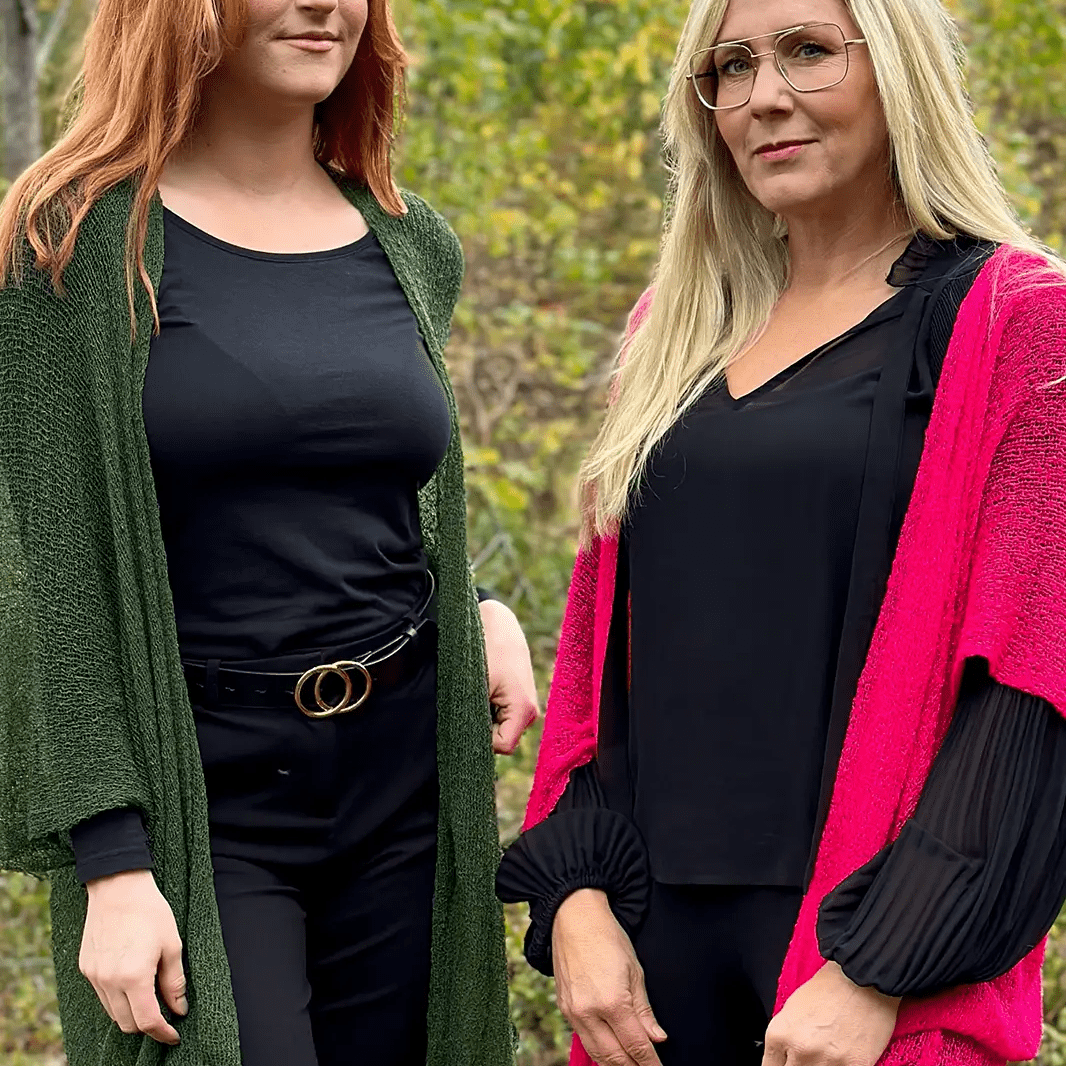 Två kvinnor sida vid sida utomhus, den ena i en mörkgrön och den andra i en neonrosa Biasa kofta – båda långa och finstickade, öppna över svarta plagg och mjukt fallande. Ulla Jacobsson Kläder