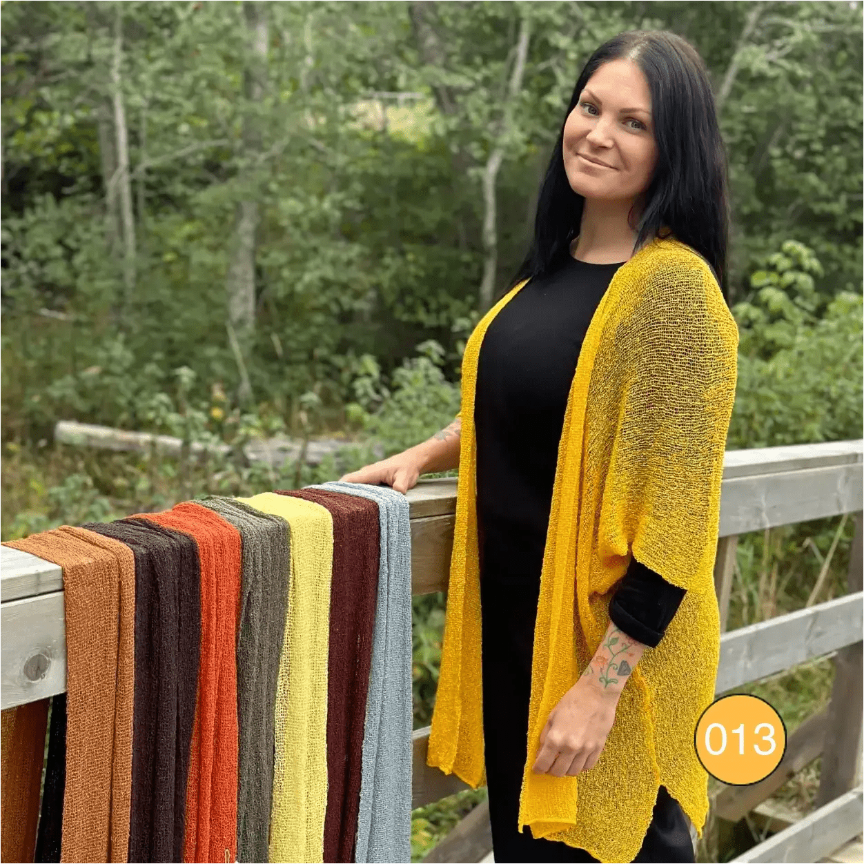 Mörkhårig kvinna utomhus vid ett räcke i en saffransgul Biasa kofta, lång och öppen över svart; det finstickade garnet faller i en varmt gul ton. Ulla Jacobsson Kläder