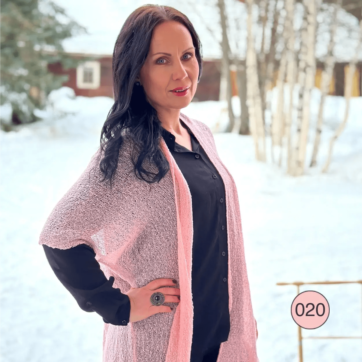 Mörkhårig kvinna i ett snötäckt vinterlandskap i en tuggummirosa Biasa kofta öppen över svart; den långa finstickade koftan lyser ljust rosa mot det vita. Ulla Jacobsson Kläder
