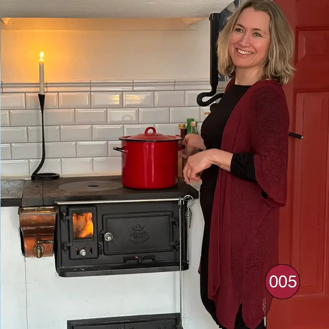 I ett kök vid en vedspis bär en kvinna en vinröd Biasa kofta löst öppen över en svart underdel; den finstickade koftan faller långt längs sidorna. Ulla Jacobsson Kläder