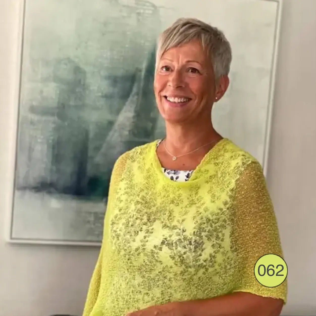 Leende äldre kvinna med kort blont hår inomhus framför en tavla, i en limegrön Ella cape vars luftiga stickning faller mjukt över axlarna. Ulla Jacobsson Kläder