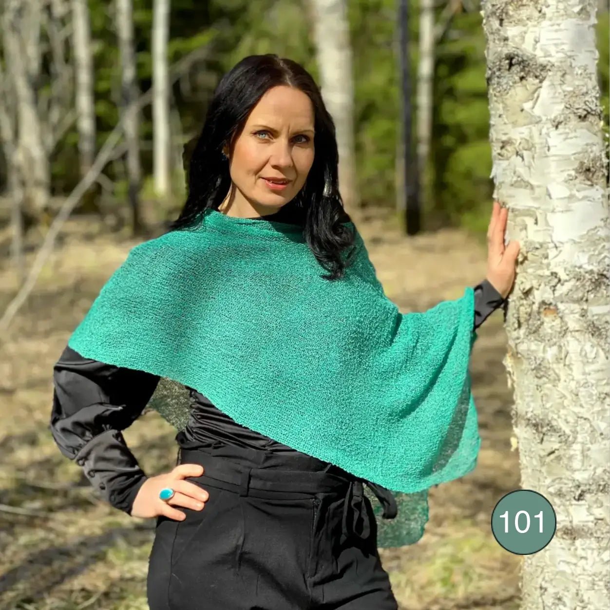 Mörkhårig kvinna med handen i sidan vid en björk, i en Ella cape i ljus buteljgrön vars luftiga stickning draperas över en svart topp. Ulla Jacobsson Kläder