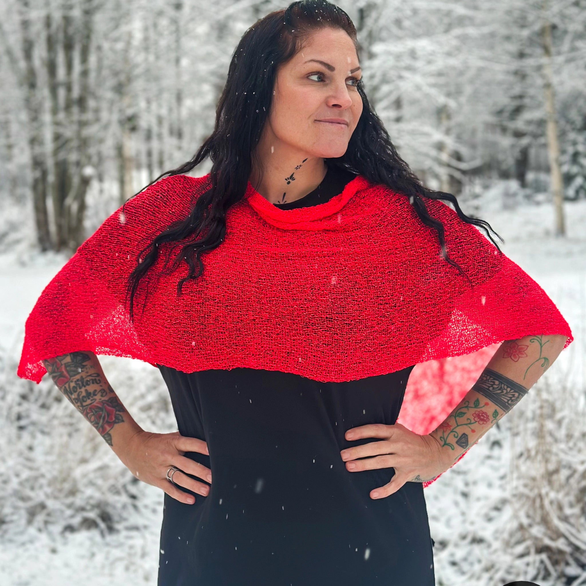 Mörkhårig kvinna med händerna i sidorna i ett snöigt skogslandskap, i en signalröd Ella cape vars skira stickning faller från axlarna över en svart klänning. Ulla Jacobsson Kläder
