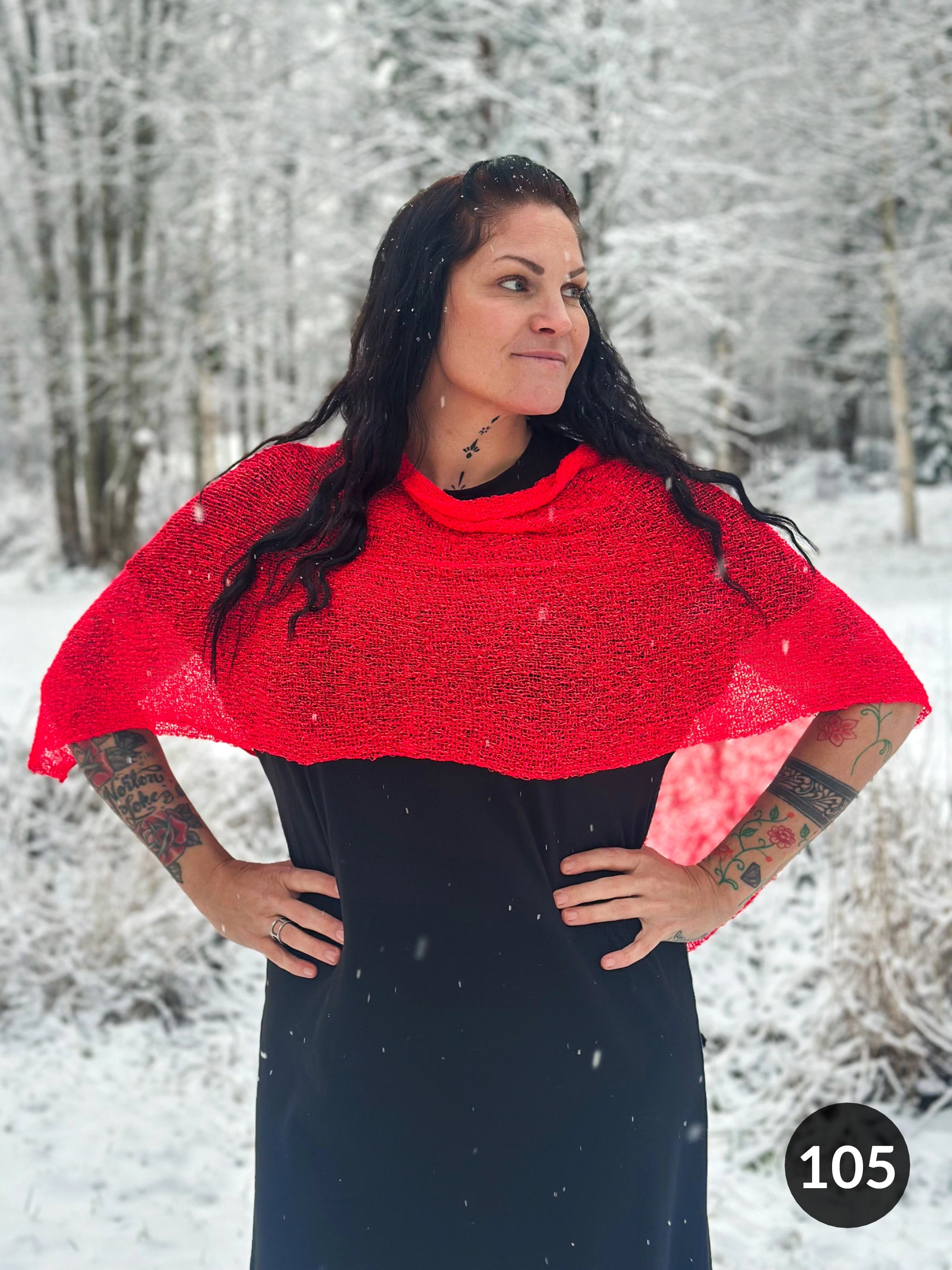 Mörkhårig kvinna med händerna i sidorna i ett snöigt skogslandskap, i en signalröd Ella cape vars skira stickning faller från axlarna över en svart klänning. Ulla Jacobsson Kläder