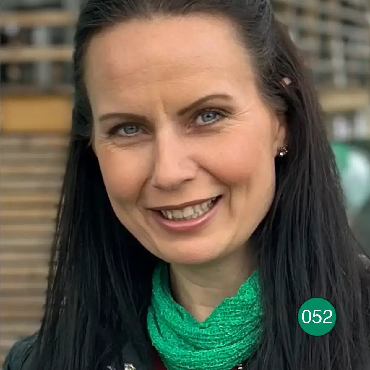 Mörkhårig kvinna som ler inomhus med en bladgrön Eva tubsjal runt halsen över svart; den lilla finstickade tuben lyser grönt. Ulla Jacobsson Kläder