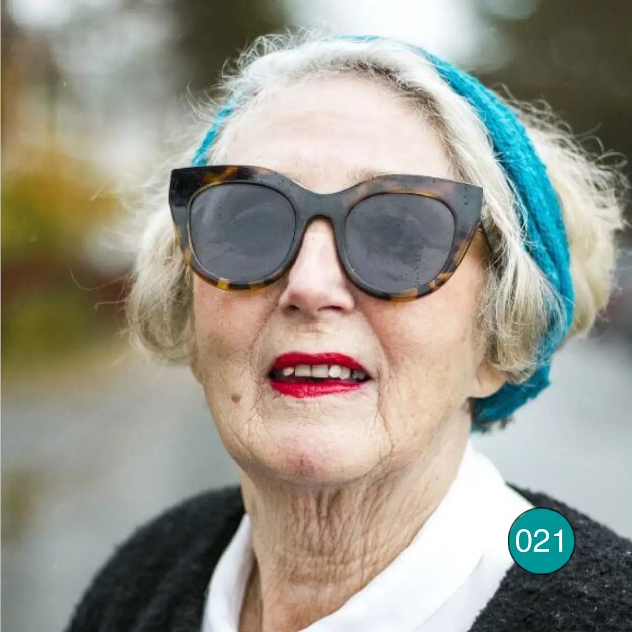 Äldre kvinna med solglasögon och grått hår bär en grönturkos Eva tubsjal som hårband; den finstickade tuben sitter brett över håret. Ulla Jacobsson Kläder