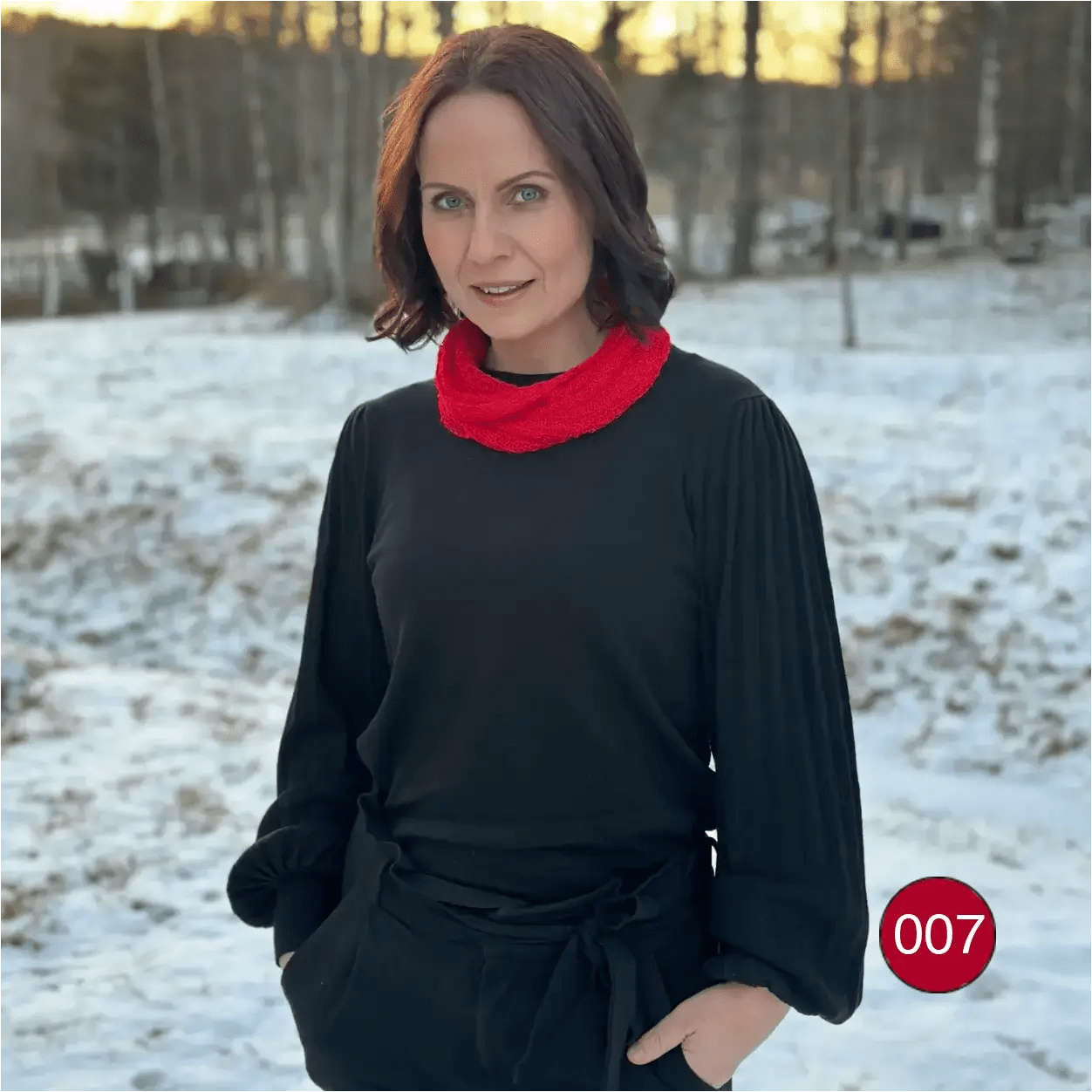 Mörkhårig kvinna med händerna i fickorna i vintermiljö, klarröd Eva tubsjal mjukt runt halsen över svart. Ulla Jacobsson Kläder