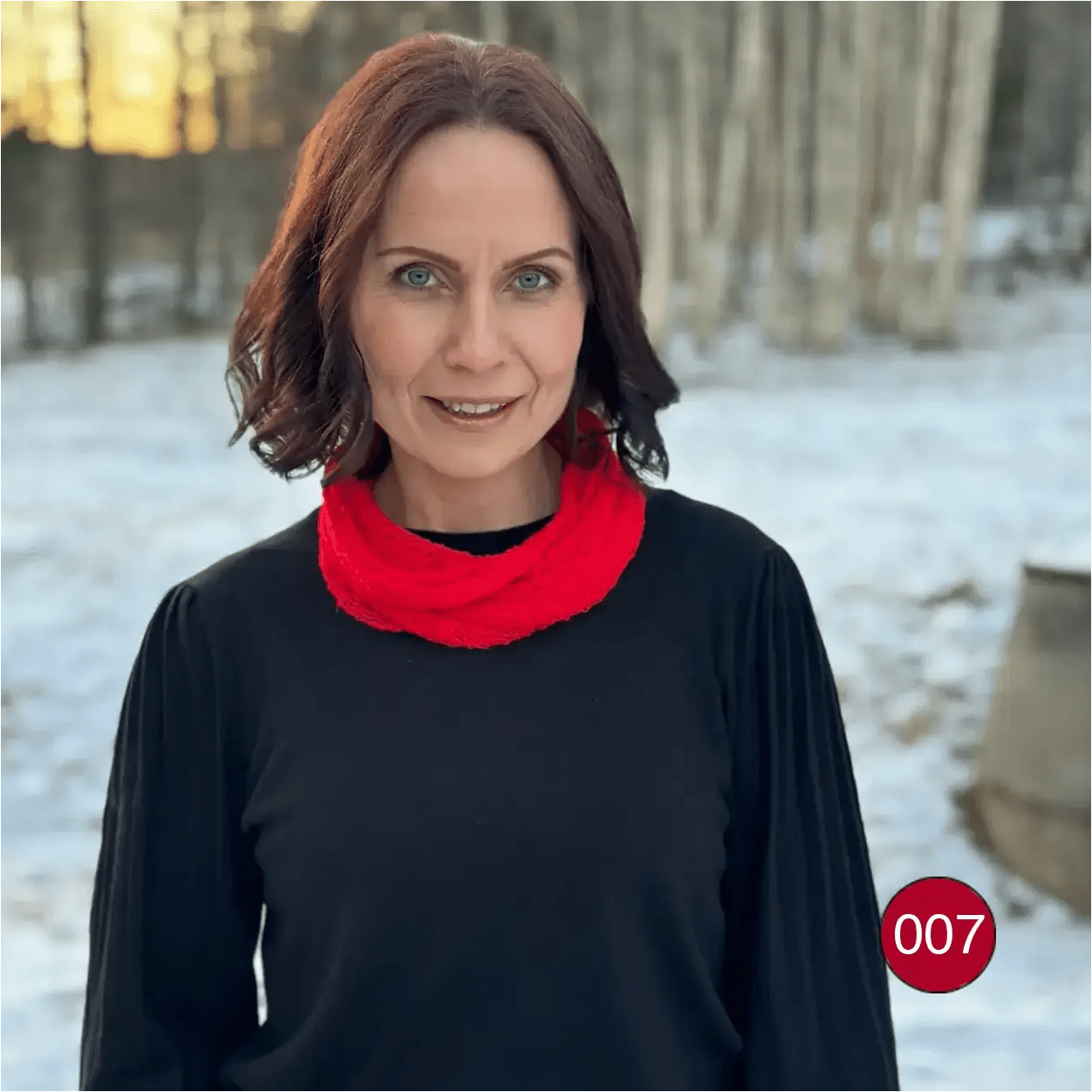 Mörkhårig kvinna utomhus med en klarröd Eva tubsjal runt halsen över en svart tröja; det finstickade garnet lyser klarrött mot snön. Ulla Jacobsson Kläder
