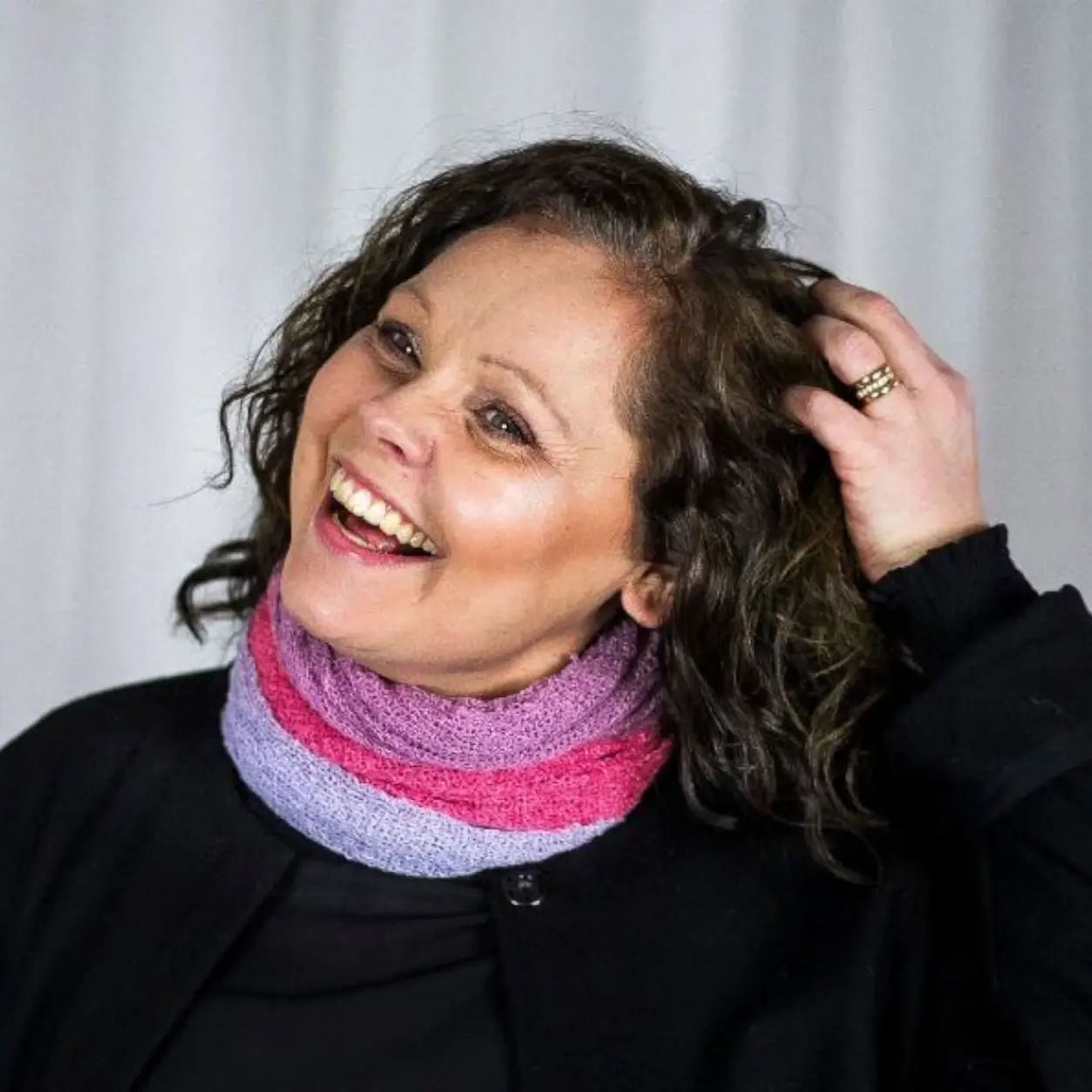 Skrattande kvinna med mörkt lockigt hår och handen i håret, flera Eva tubsjalar lagrade runt halsen i lavendel, cerise och lila över svart. Ulla Jacobsson Kläder