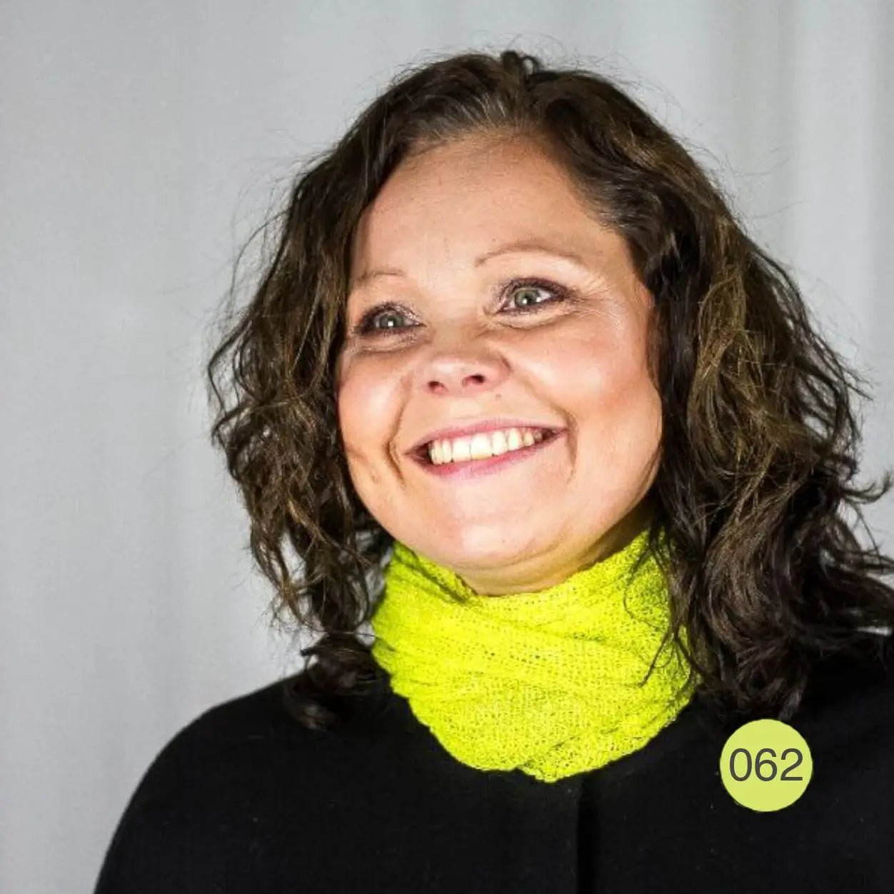 Kvinna med lockigt hår och ett brett leende, en limegrön Eva tubsjal runt halsen över svart; det finstickade garnet lyser gulgrönt. Ulla Jacobsson Kläder