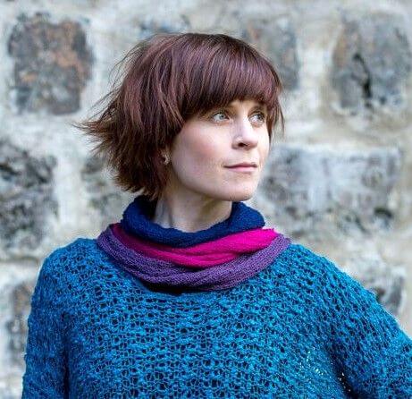 Korthårig kvinna mot en stenvägg med flera Eva tubsjalar lagrade runt halsen i marinblått, cerise och lila, buren till en blå stickad tröja. Ulla Jacobsson Kläder