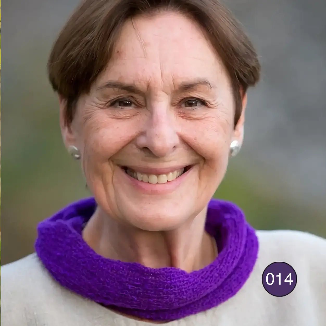 Äldre kvinna med kort hår ler mot kameran med en mörklila Eva tubsjal mjukt vikt runt halsen; den lilla, finstickade tubsjalen ligger len mot huden. Ulla Jacobsson Kläder