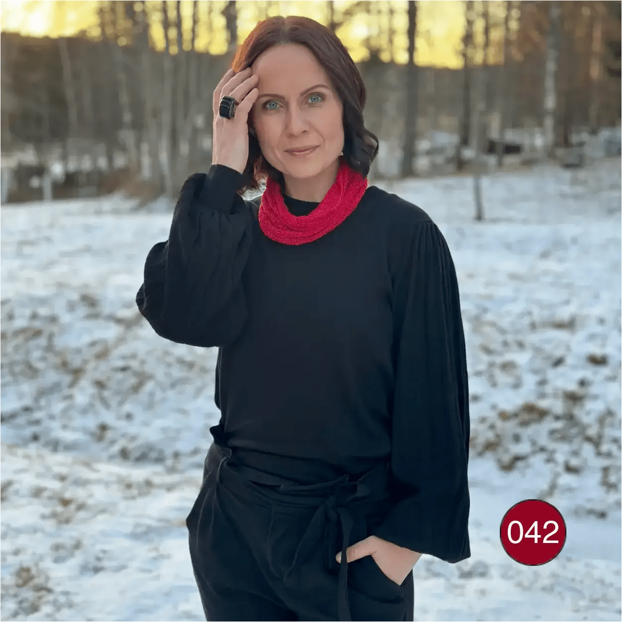 Mörkhårig kvinna i snölandskap som för handen mot håret, röd Eva tubsjal mjukt vikt runt halsen över svart. Ulla Jacobsson Kläder