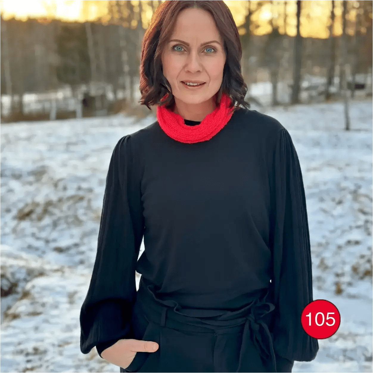 Mörkhårig kvinna i snölandskap med en signalröd Eva tubsjal mjukt vikt runt halsen över svart. Ulla Jacobsson Kläder