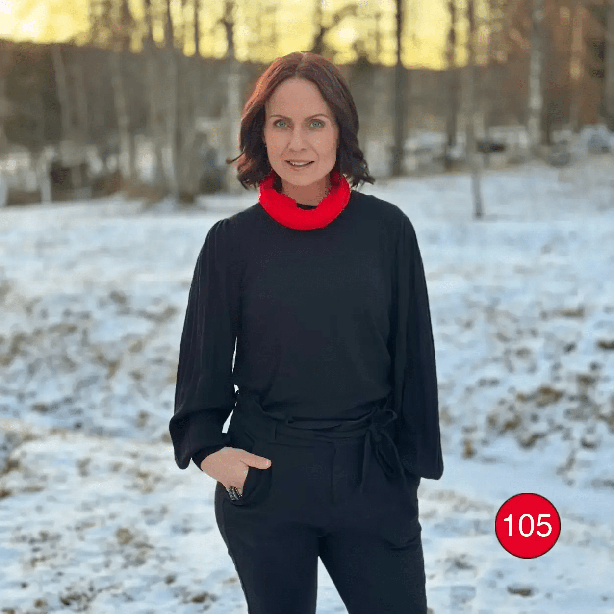 Mörkhårig kvinna utomhus i vintermiljö med en signalröd Eva tubsjal runt halsen över en svart tröja. Ulla Jacobsson Kläder