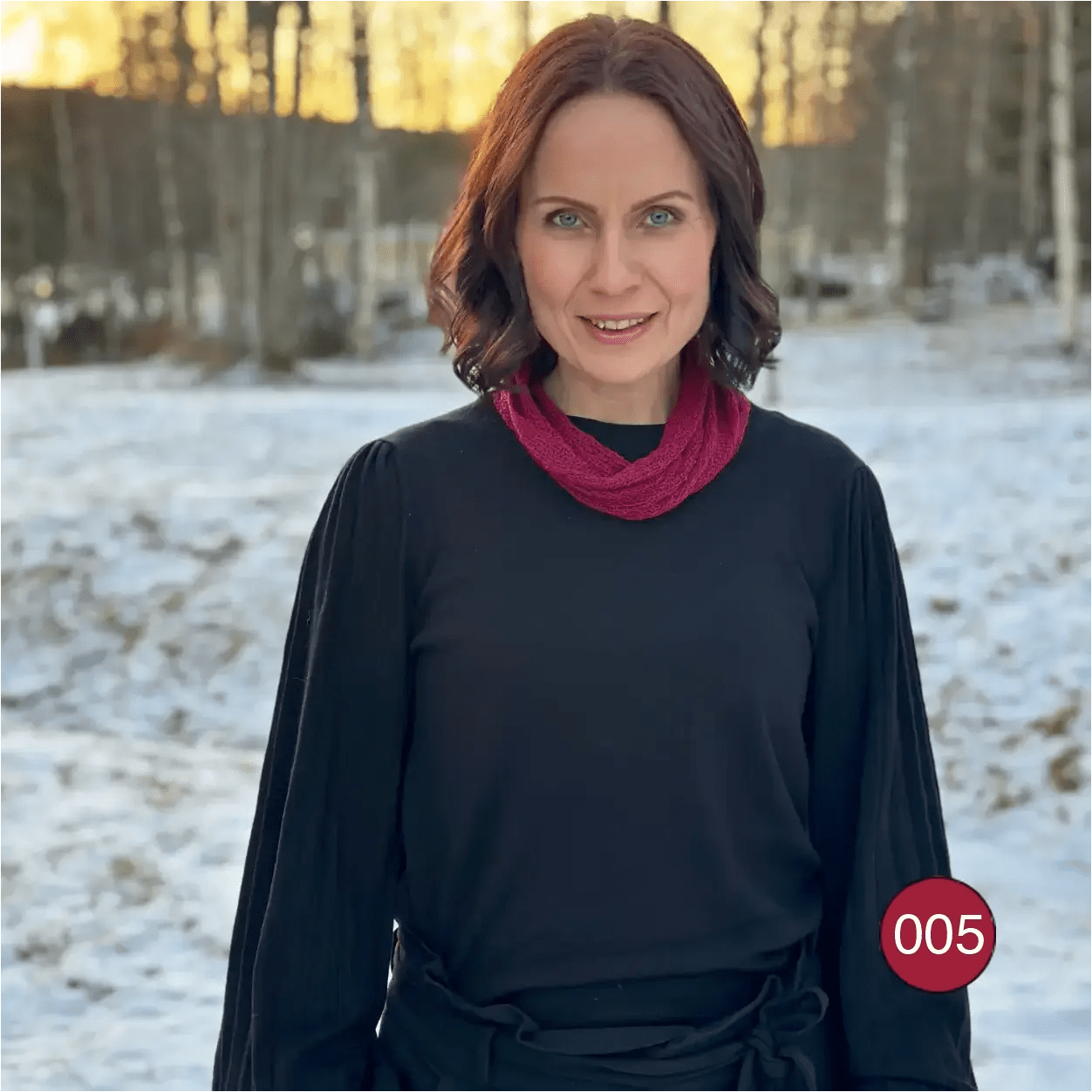 Mörkhårig kvinna i snölandskap, vinröd Eva tubsjal lagd dubbelt runt halsen över svart; det finstickade garnet ramar in ansiktet. Ulla Jacobsson Kläder