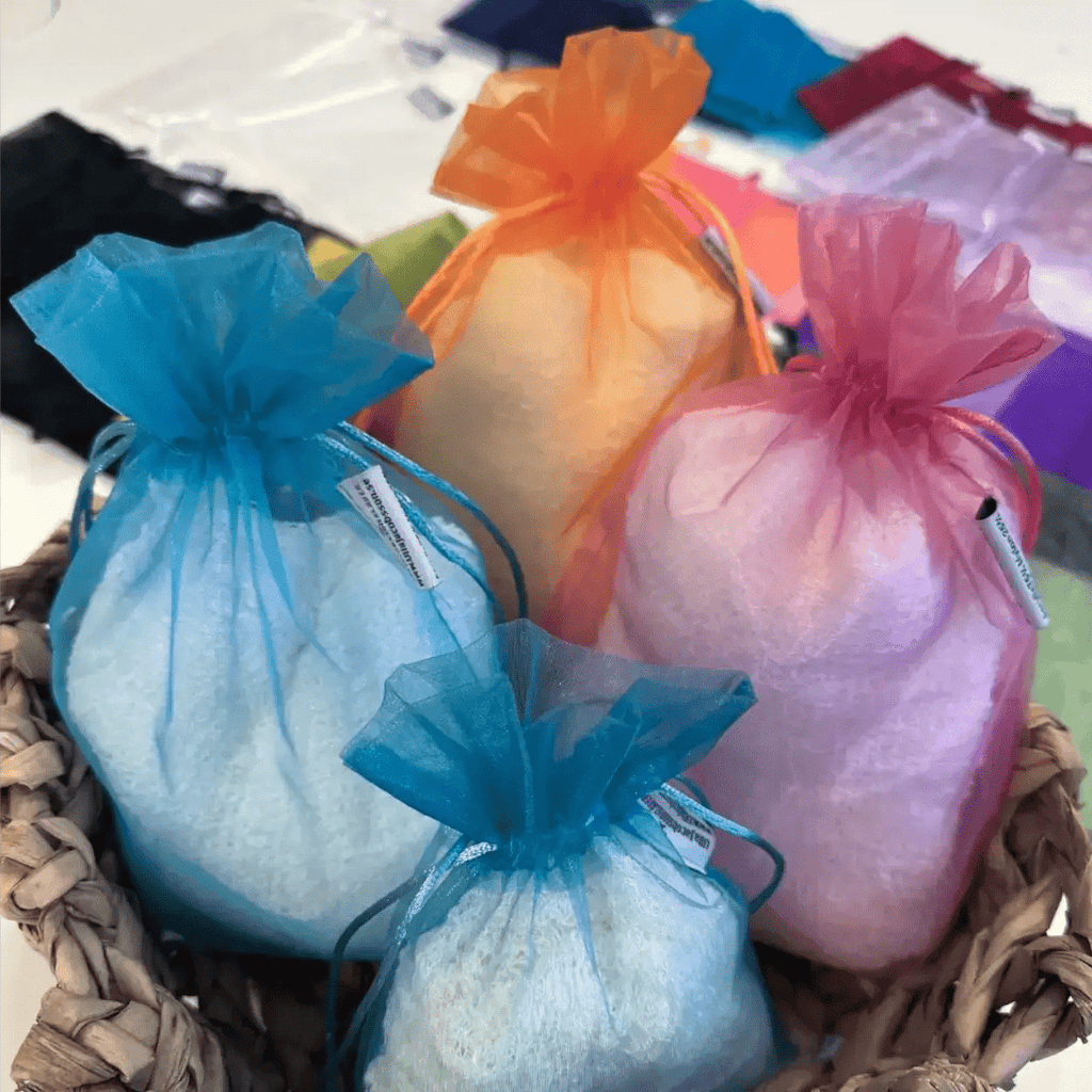Färgglada förvaringspåsar i organza i turkos, rosa, lila och orange, fyllda med finstickade plagg och samlade på ett ljust underlag. De tunna organzapåsarna låter plaggen andas samtidigt som de skyddas vid förvaring. Ulla Jacobsson Kläder
