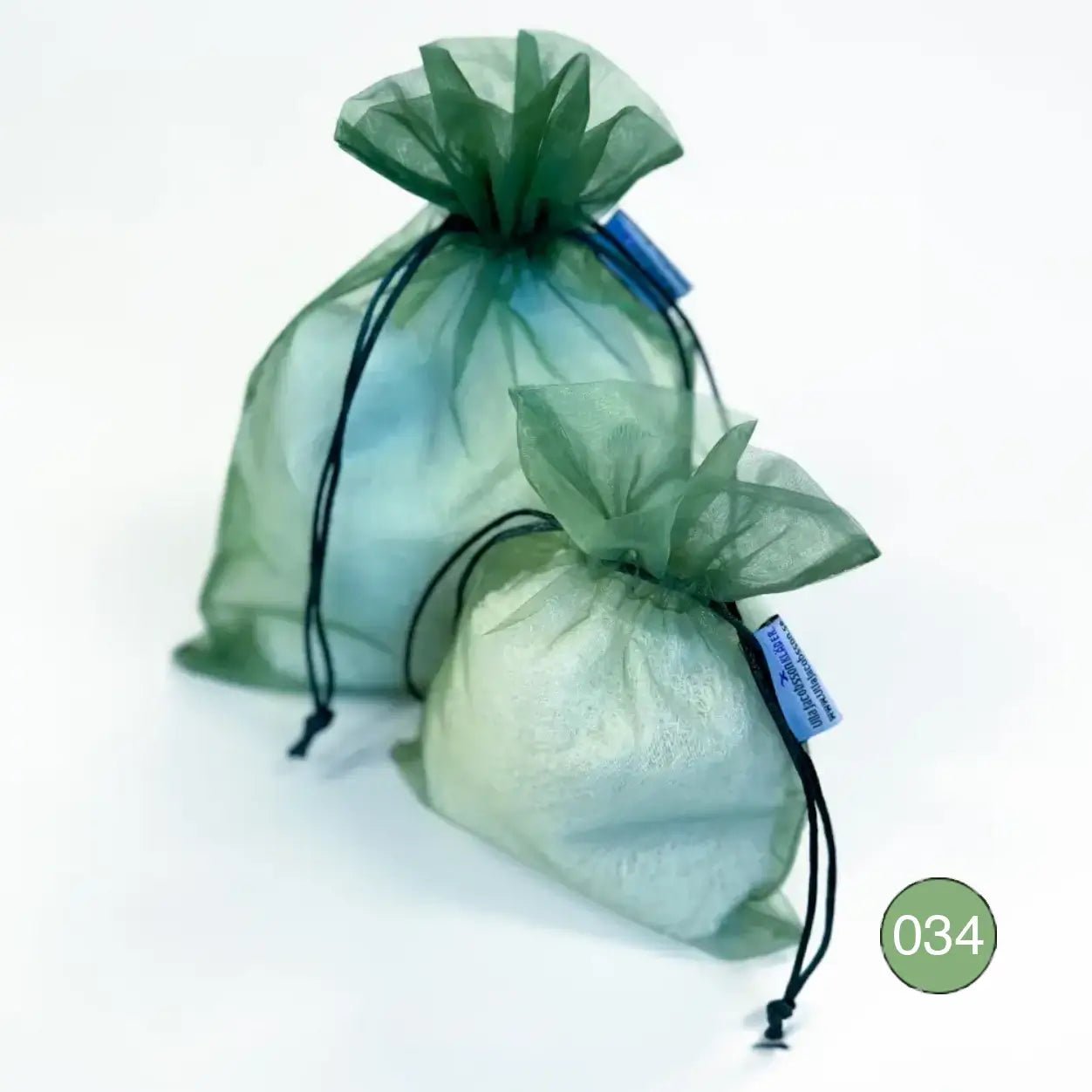 Kaksi vaaleanvihreää organzaista säilytyspussia, pieni ja suurempi, täytettyinä vaalealla ohutneulotulla vaatteella valkoisella taustalla. Ohut organza antaa vaatteen hengittää ja suljetaan nyörellä. Ulla Jacobsson Kläder