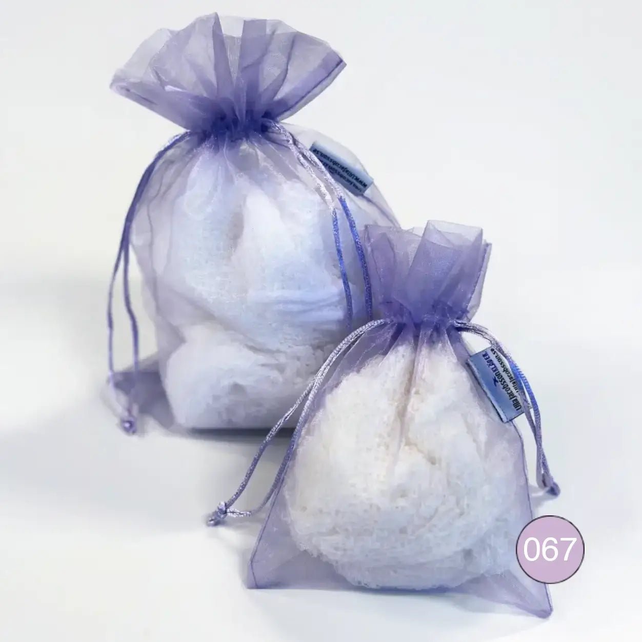 Kaksi vaaleanviolettia organzaista säilytyspussia, pienempi ja suurempi, täytettyinä vaalealla ohutneulotulla vaatteella valkoisella taustalla. Ohut organza hengittää ja suljetaan nyörellä. Ulla Jacobsson Kläder