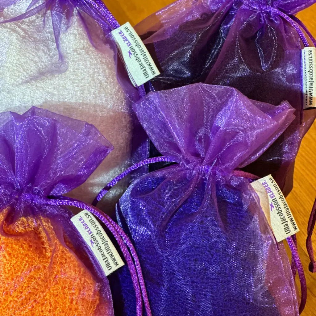 Lähikuva tummanvioleteista organzaisista säilytyspusseista, jotka on täytetty ohutneulotuilla vaatteilla ja suljettu nyörellä; pussin kyljessä on pieni kirjailtu logo. Ulla Jacobsson Kläder