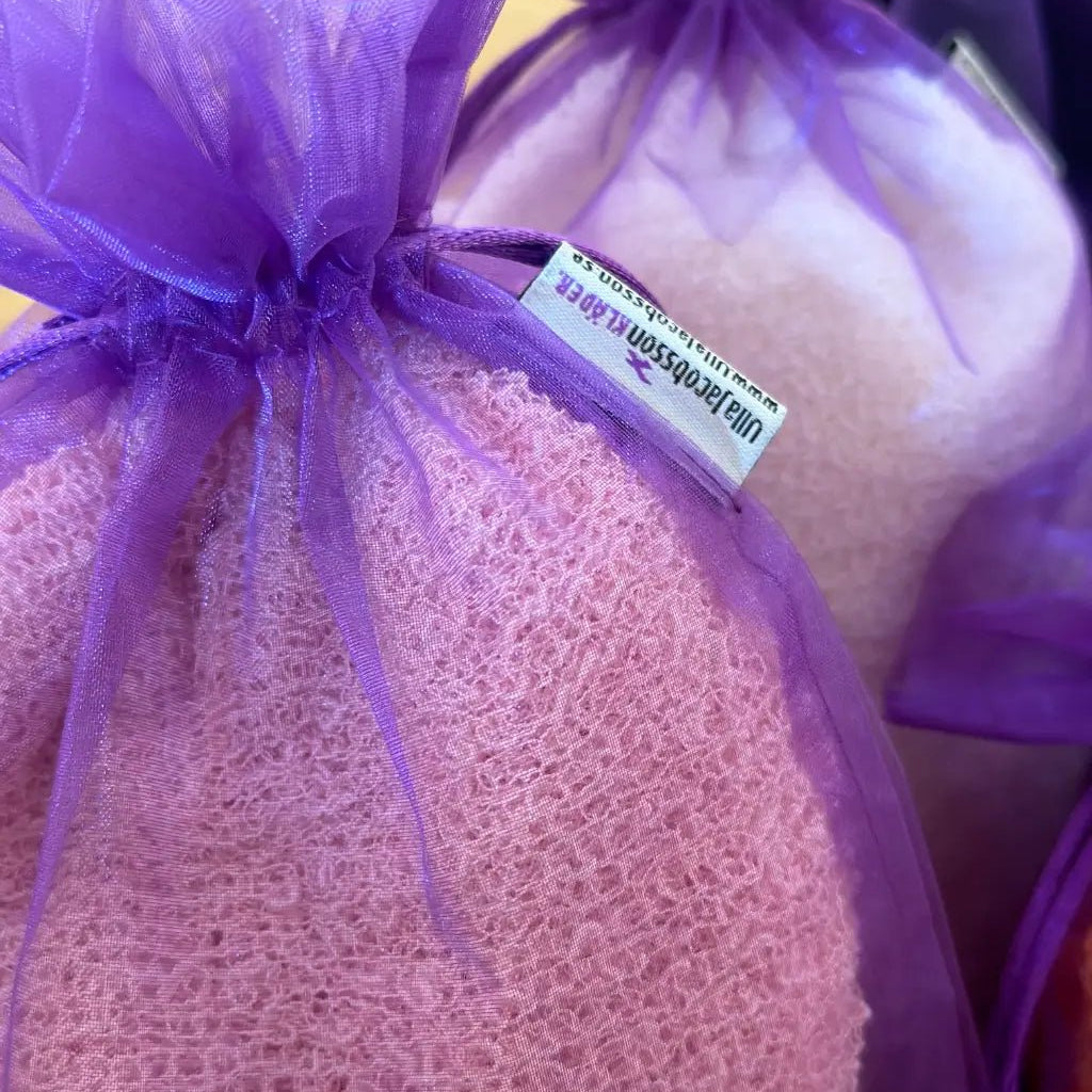 Lähikuva tummanvioletista organzapussista, joka on täytetty roosalla ohutneulotulla vaatteella; läpikuultava kangas näyttää vaatteen ja suojaa sitä pölyltä ja kulumiselta. Ulla Jacobsson Kläder