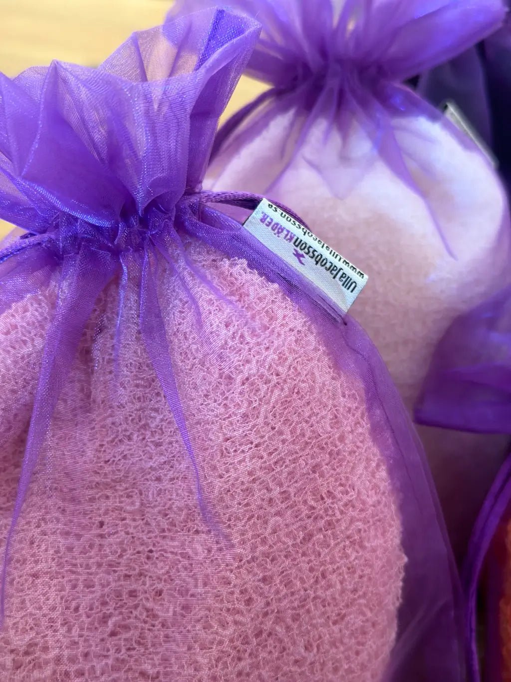 Lähikuva tummanvioletista organzapussista, joka on täytetty roosalla ohutneulotulla vaatteella; läpikuultava kangas näyttää vaatteen ja suojaa sitä pölyltä ja kulumiselta. Ulla Jacobsson Kläder