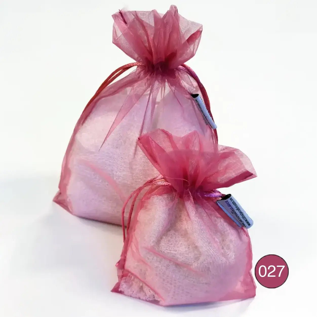 Kaksi roosaa organzaista säilytyspussia, pienempi ja suurempi, täytettyinä vaalealla ohutneulotulla vaatteella valkoisella taustalla. Ohut organza hengittää ja suljetaan nyörellä. Ulla Jacobsson Kläder