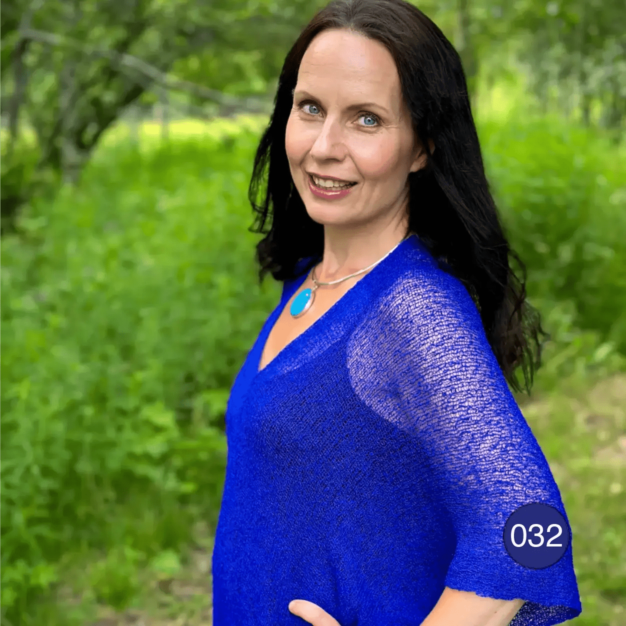 Leende mörkhårig kvinna i halvbild med ett turkost halssmycke, i en klarblå Idun tunika; det finstickade garnet syns tunt och luftigt och faller mjukt över axlarna mot den gröna bakgrunden. Ulla Jacobsson Kläder