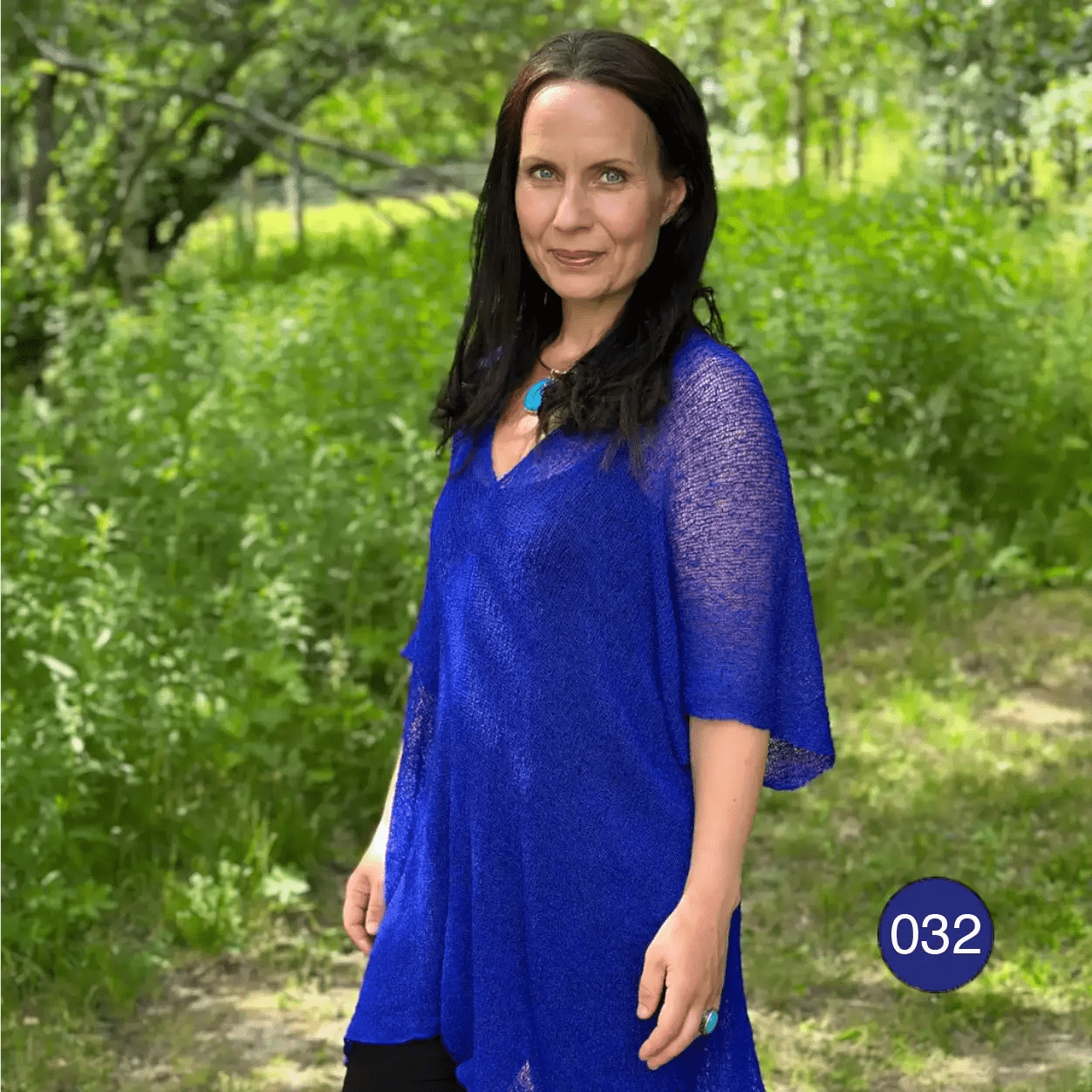 Mörkhårig kvinna på en grön stig i en klarblå Idun tunika med trekvartsärm; den finstickade oversize-tunikan faller mjukt över axlar och armar i en lätt, luftig siluett. Ulla Jacobsson Kläder
