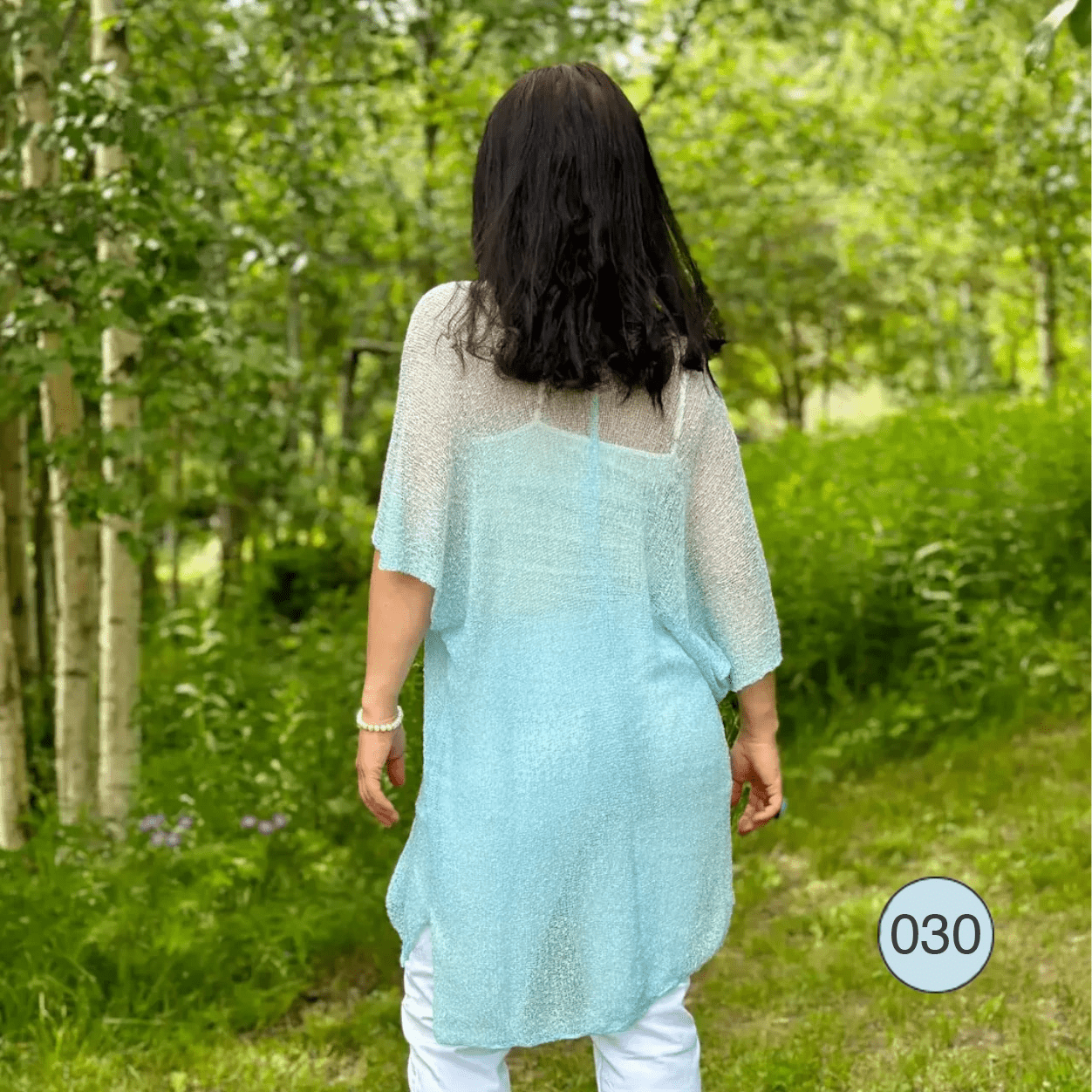 Kvinna fotograferad bakifrån i björkskogen visar ryggen på en turkosblå Idun tunika; det luftiga finstickade garnet faller mjukt ned över vita byxor i en lätt, knälång siluett. Ulla Jacobsson Kläder