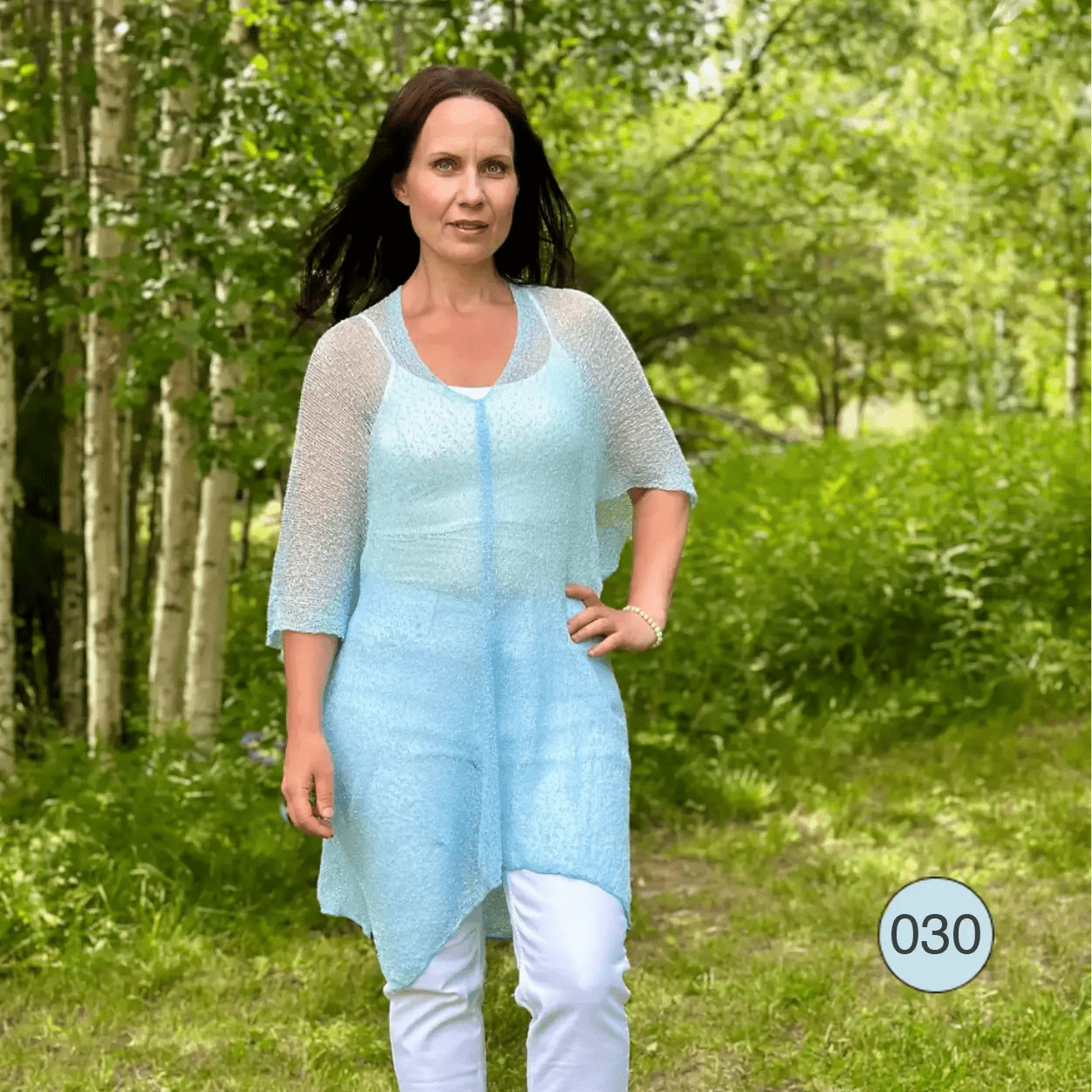 Mörkhårig kvinna med handen i sidan i en turkosblå Idun tunika med trekvartsärm, buren över vita byxor i sommargrön björkskog; det finstickade garnet är tunt och luftigt. Ulla Jacobsson Kläder