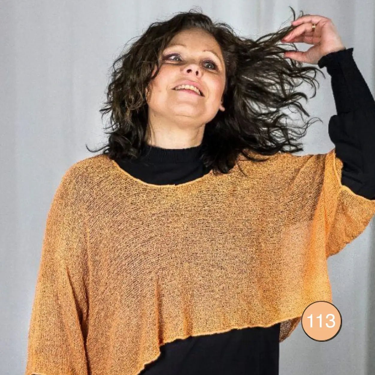 Mörkhårig kvinna med håret i rörelse i en aprikosfärgad Lebar poncho; det finstickade garnet i varm gyllene ton faller draperat över en svart underdel i ett luftigt fall. Ulla Jacobsson Kläder