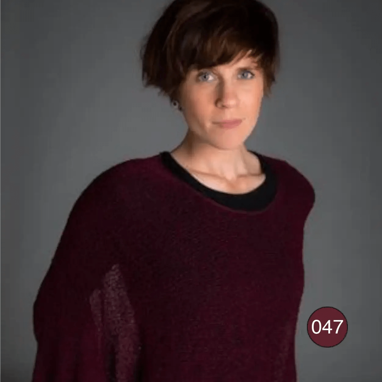Korthårig kvinna i en bourdeauxröd Lebar poncho mot mörk bakgrund; det tunna finstickade garnet faller mjukt och draperat över en svart topp i en djup vinröd ton. Ulla Jacobsson Kläder