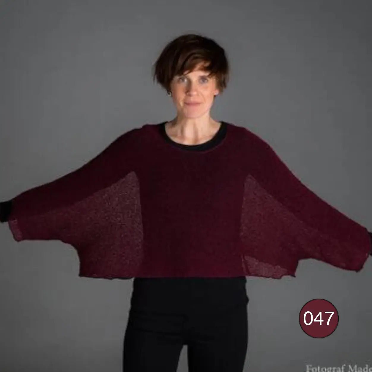 Korthårig kvinna sträcker ut armarna i en bourdeauxröd Lebar poncho så den vida finstickade ärmen breder ut sig; den tunna ponchon bärs över svart och visar sitt luftiga fall. Ulla Jacobsson Kläder