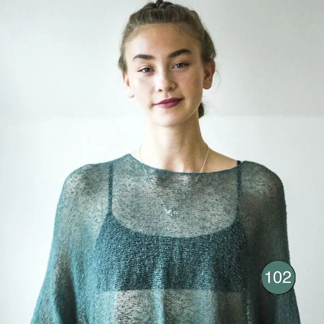 Ljushårig kvinna med uppsatt hår i en buteljgrön Lebar poncho; det tunna, nästan genomskinliga finstickade garnet faller mjukt i djup grönblå ton och draperar löst över axlarna. Ulla Jacobsson Kläder