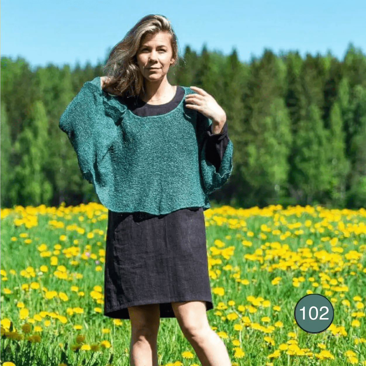 Ljushårig kvinna i en sommaräng bär en buteljgrön Lebar poncho över en mörk kjol; den finstickade ponchon med vida ärmar draperar löst över axlar och armar. Ulla Jacobsson Kläder