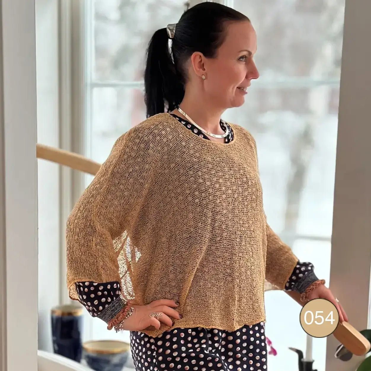 Mörkhårig kvinna med uppsatt hår och handen i sidan i en kamelpärgad Lebar poncho inomhus; det finstickade garnet i varm beigebrun ton ligger luftigt och faller mjukt över axlarna. Ulla Jacobsson Kläder